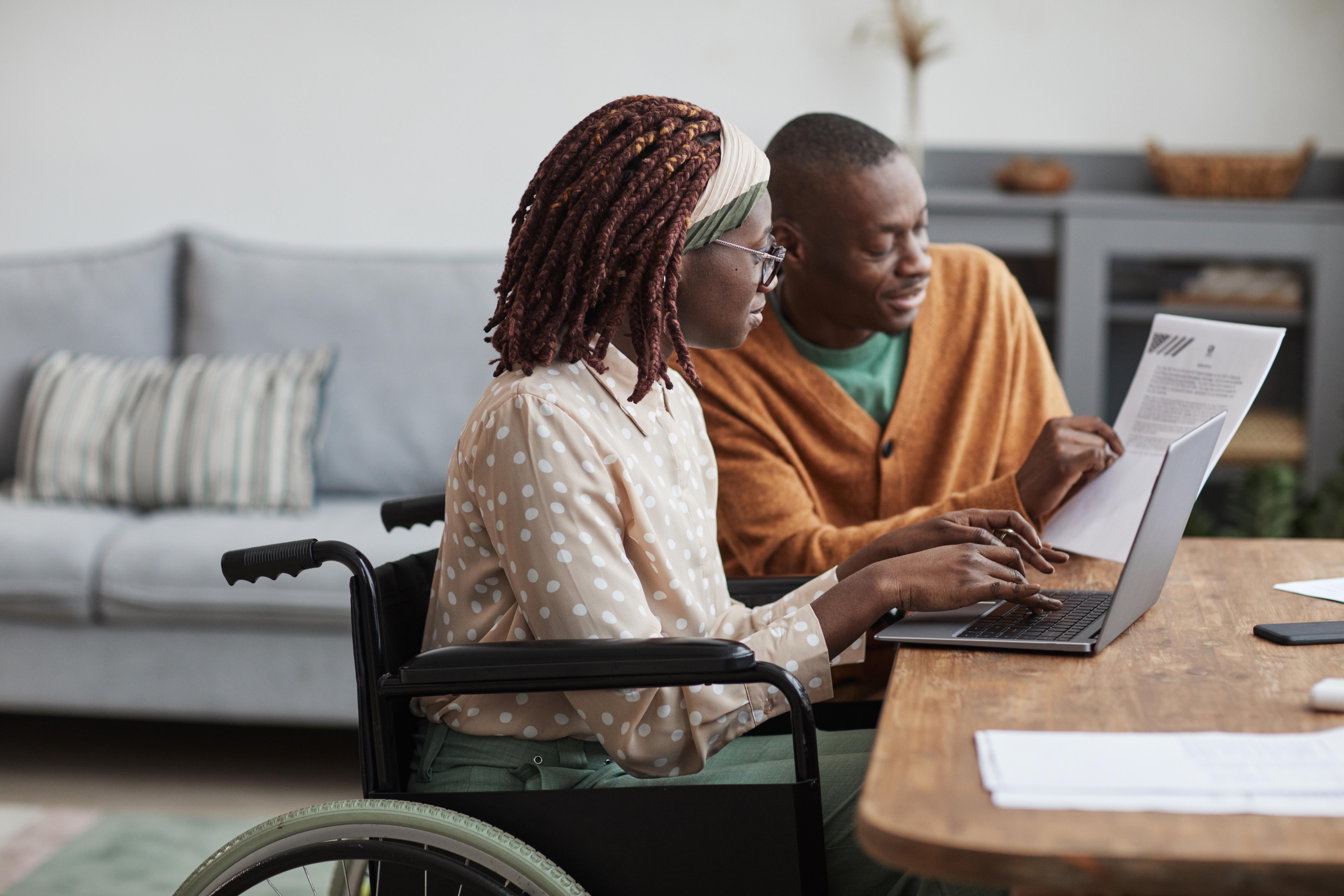 Woman in wheel chair on laptop with man assisting